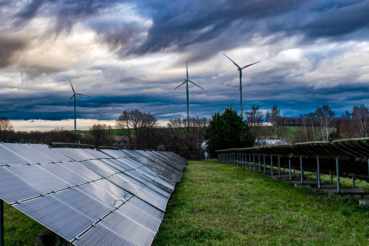 Adani Group Q2 results: rows of solar panels with wind turbines behind them under a cloudy sky