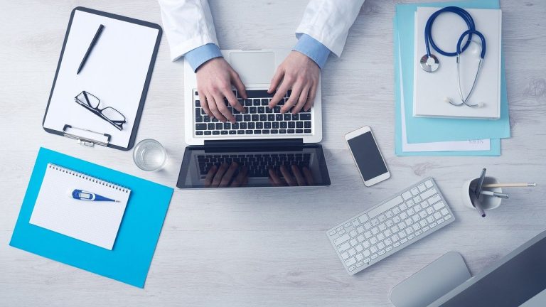 Sagility Chirok Health acquisition: doctor typing on a laptop at a desk with medical paperwork
