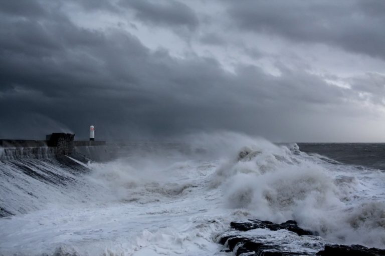 Implied volatility vs historical volatility: storm waves crashing over a harbour wall with a small lighthouse under dark clouds
