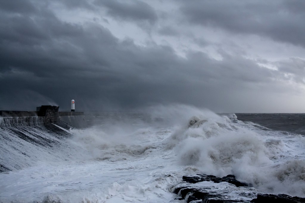 Implied volatility vs historical volatility: storm waves crashing over a harbour wall with a small lighthouse under dark clouds