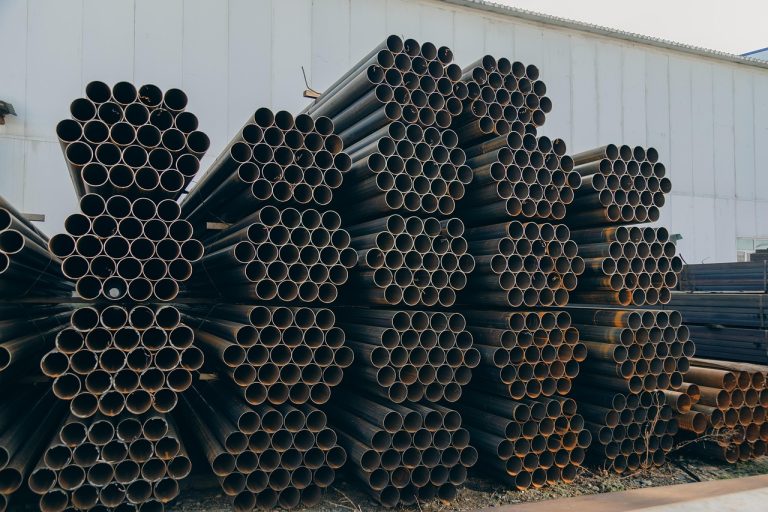 Jindal Saw Q2 results: stacks of black steel pipes stored outside a factory shed